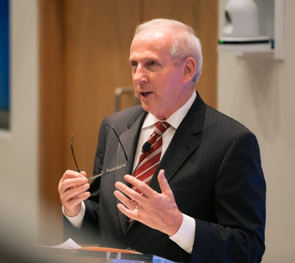 Man speaking at podium holding eyeglasses