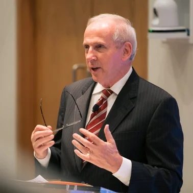 Man speaking at podium holding eyeglasses